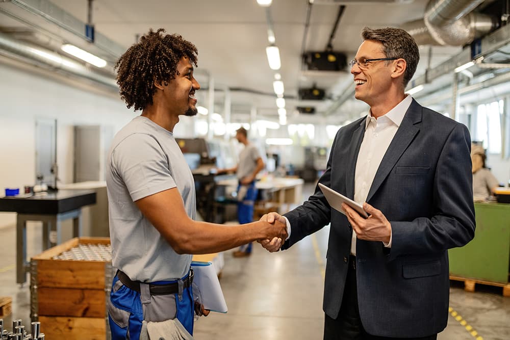 Worker and manager shaking hands in a manufacturing facility, one holding a tablet.