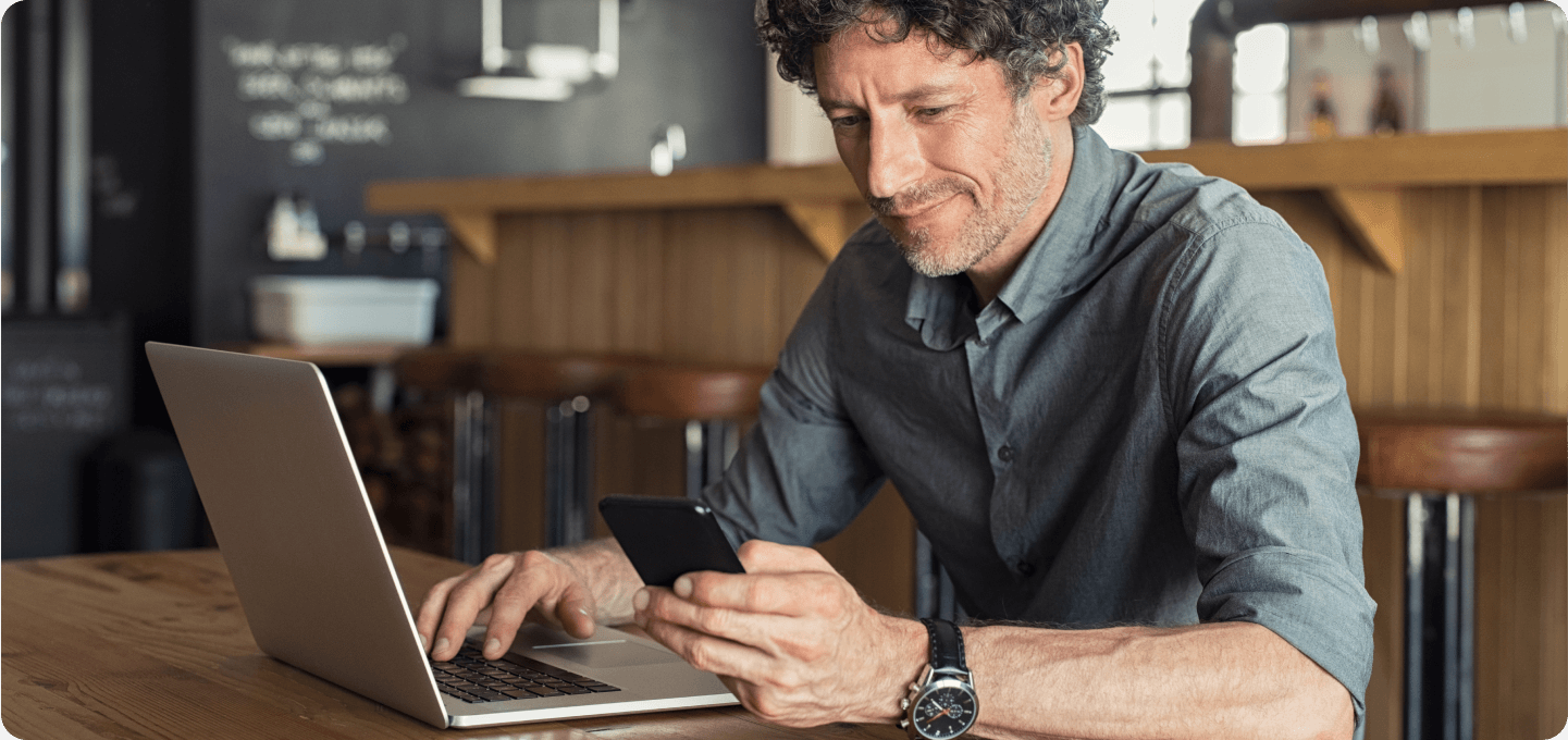 Middle-aged man using a smartphone while working on a laptop in a café.