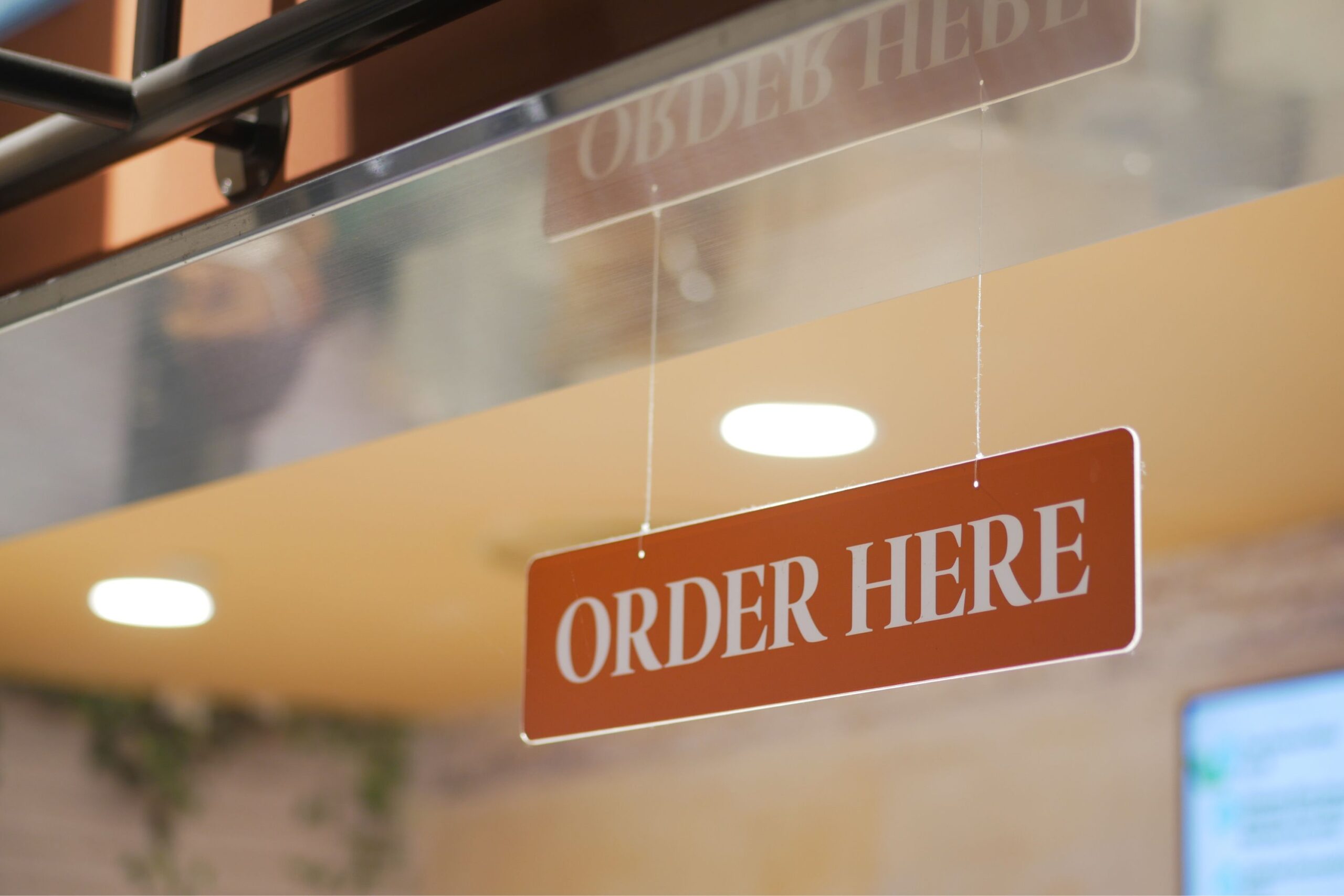 Orange hanging sign reading 'ORDER HERE' near a glass counter in a cafe/restaurant setting.