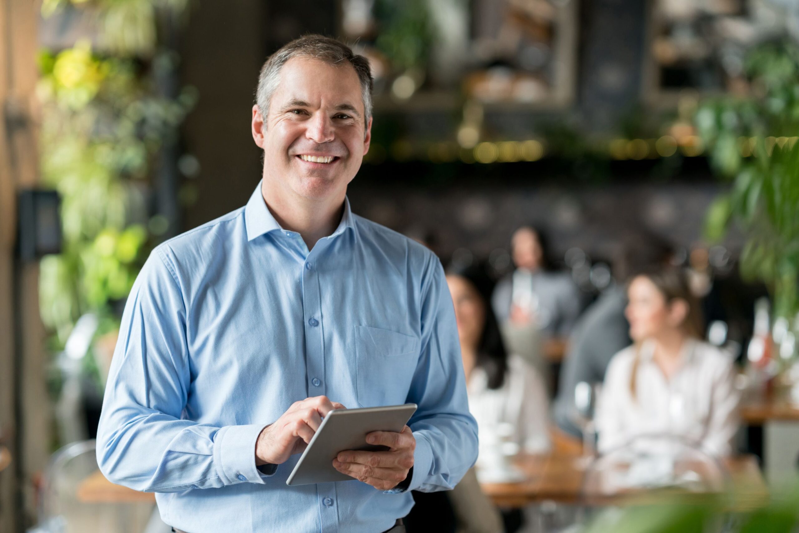Actuate – Act Hub 2.0 Operational Intelligence Smiling man in a light blue shirt holding a tablet in a busy cafe setting.