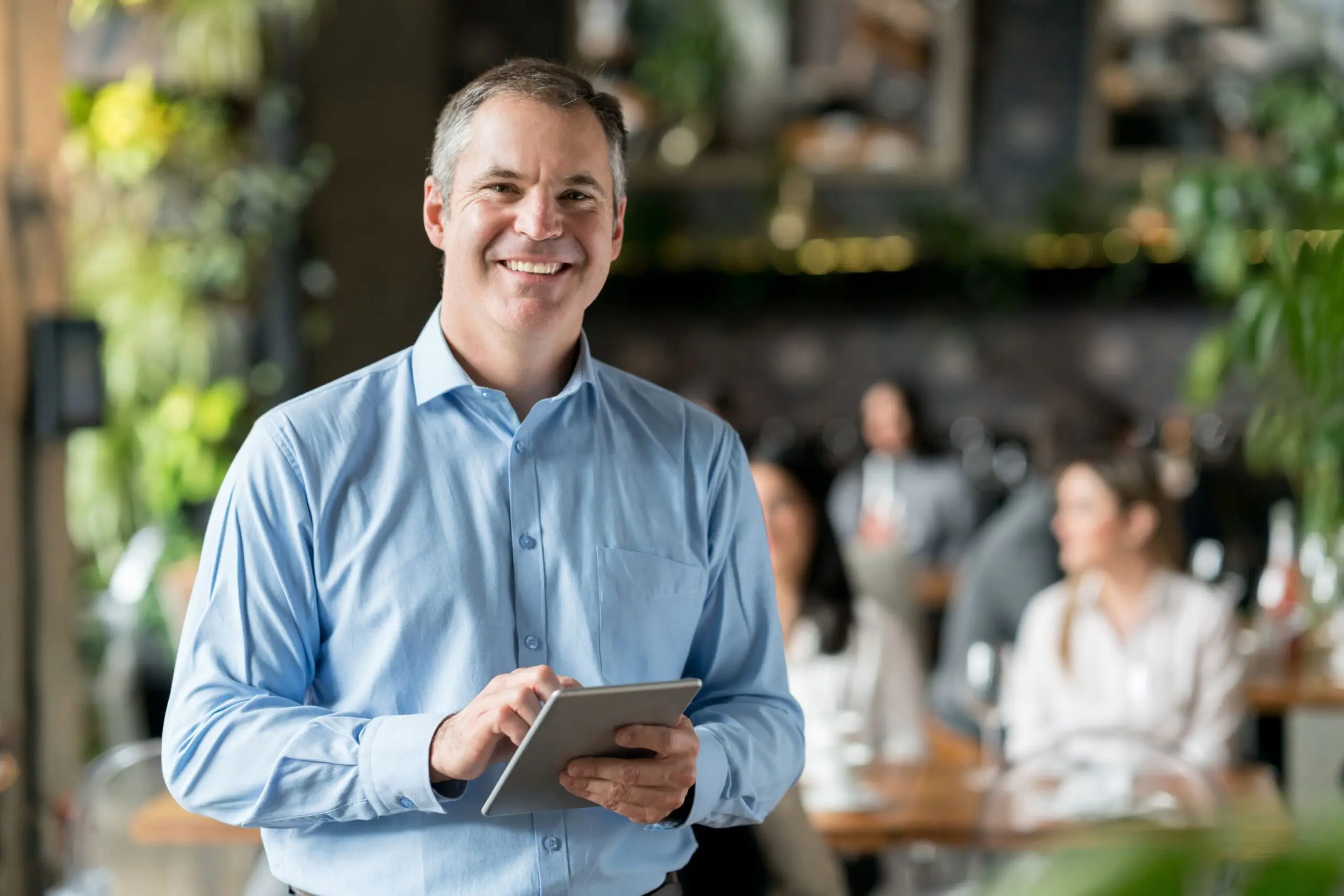 Smiling man in a light blue shirt holding a tablet in a busy cafe setting.