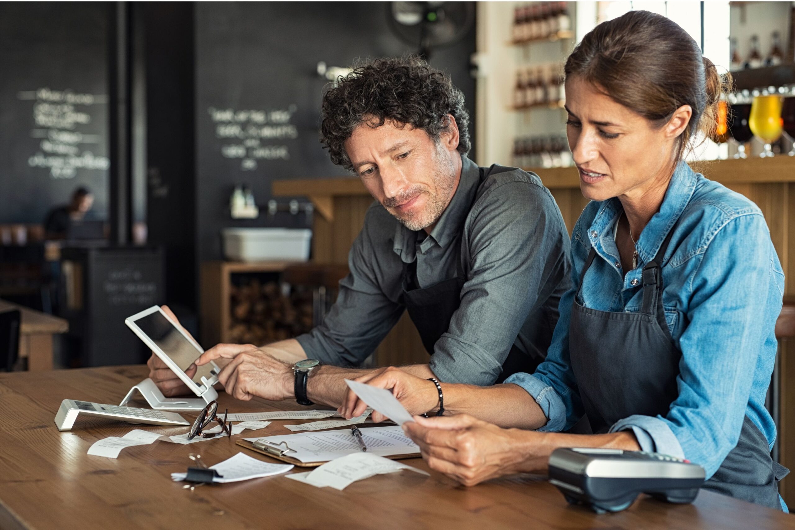 Two coworkers in aprons review receipts at a cafe counter, using a tablet and calculator.