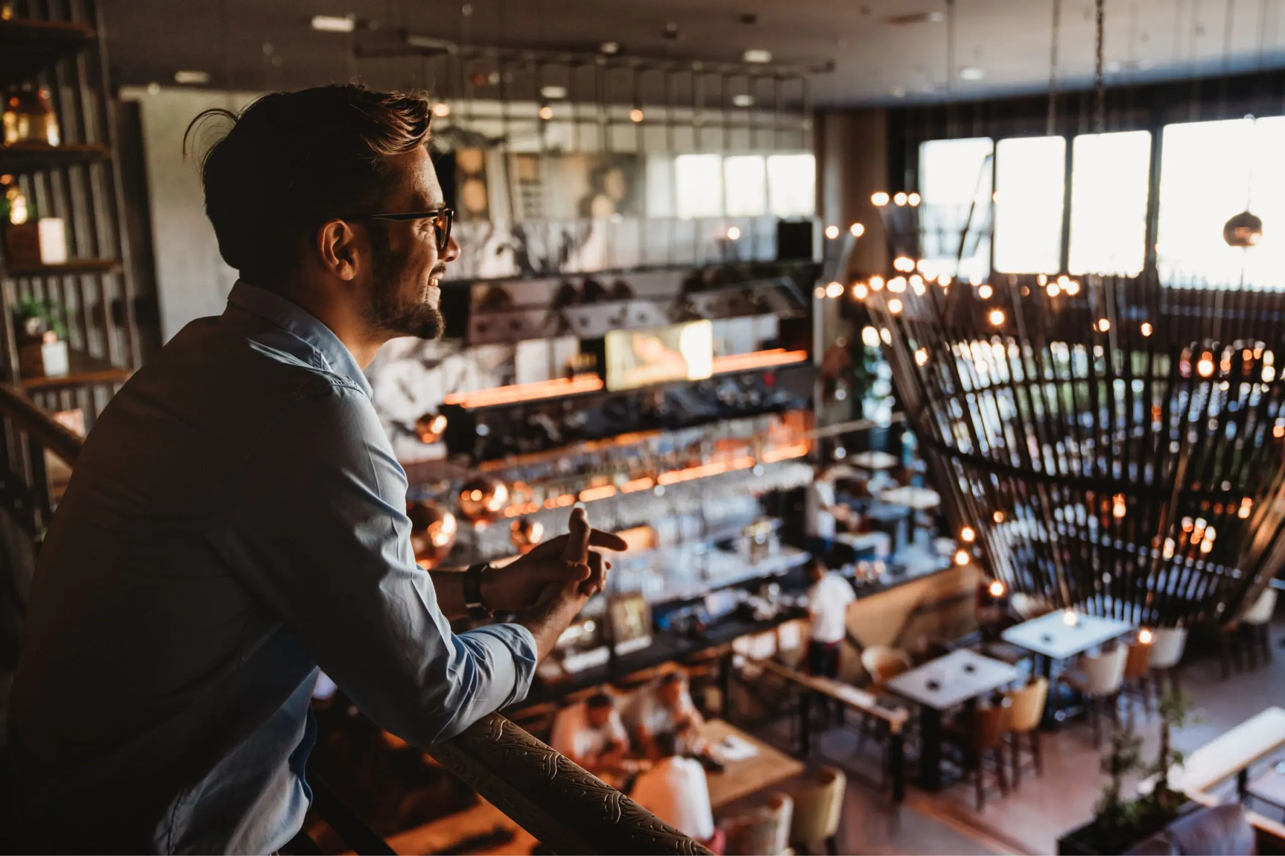Man with glasses leaning on a railing, smiling in a busy, warmly lit restaurant or cafe mezzanine.